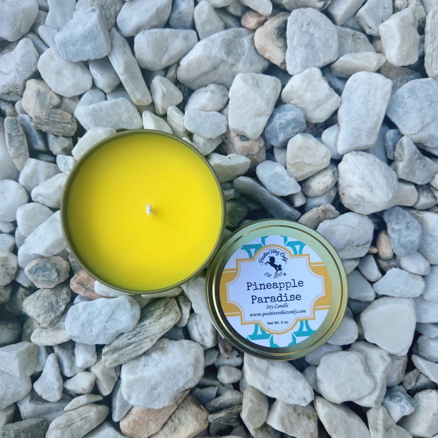 Yellow candle in a tin labeled 'Pineapple Paradise' on pebbles